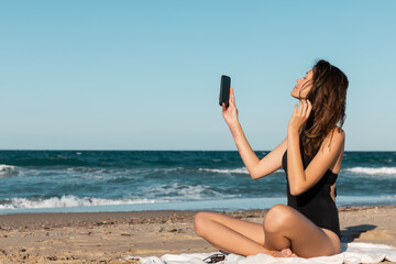 Wall Mural - brunette young woman in swimsuit taking selfie while sitting on blanket near sea.