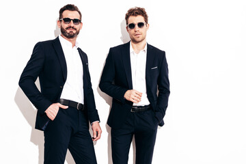 Portrait of two handsome confident stylish hipster lambersexual models. Sexy modern men dressed in black elegant suit. Fashion male posing in studio near white wall. In sunglasses