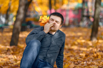 portrait of a man sitting in a glade with yellow maple leaves and scratching his nose, allergy concept, bright sunny day in an autumn park