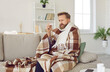 © Studio Romantic - Sick bearded man who has bad cold or seasonal flu sitting on couch at home. Guy with fever wearing warm scarf and plaid shivering and looking at thermometer in hand with worried face expression