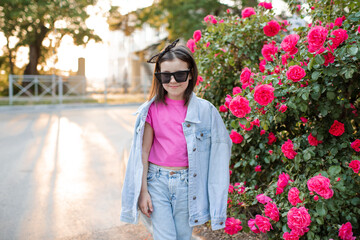 Wall Mural - Smiling cute kid girl 6-7 year old wear mom denim jacket and sun glasses posing over pink rose flowers over nature background. Childhood. Summer season.