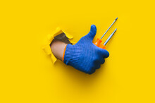 A Right Hand In A Blue Cloth Knitted Glove Holds A Two Orange Screwdriver. Torn Hole In Yellow Paper. The Concept Of A Worker, Labor Migrant, A Master Of His Craft. Copy Space.