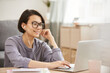 © Mediaphotos - Cheerful attractive young businesswoman in eyeglasses wearing earphones sitting at table and using laptop while calling on video conferencing app