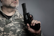A Man With A Gray Beard And In Camouflage Blows On The Barrel Of A Pistol. Military Conflicts, Murders And Crimes. Grey Background. Close-up.