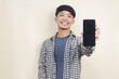 Close-up portrait of happy young Asian male model wearing plaid shirt pointing at phone screen on isolated background