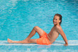 © servando - 10 year old blond boy is lying in the pool in the water, wearing orange swimming trunks.