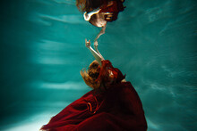 Photo Underwater, A Young Beautiful Woman In Red With Red Hair Reaches For The Surface Of The Water, A Human And His Reflection. Mystical Underwater Portrait