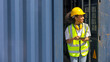 © Ron - Young African American female trainee uses tablet to check cargo containers and stocks in a logistic warehouse. Female worker in hardhat, earmuff, and safety suit stands next to a blue container.
