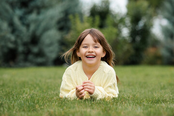 Funny smiling blonde little child girl 4-5 year old lying on green grass lawn outdoor. Small kid wear knitted casual sweater over nature background in park. Childhood. Autumn season.