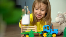 Close Up Of Toy Tractor Delivers Milk In Cart To Little Girl Lying On Floor. Happy Child Laughs And Drinks Milk From Bottle With Straw