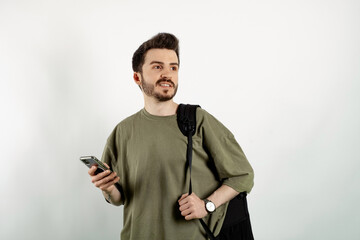 Wall Mural - Handsome young man wearing casual clothes posing isolated over white background looking far away to the front while holding a mobile phone.