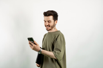 Wall Mural - Young man wearing khaki t-shirt posing isolated over white background holding in hands carrying laptop and phone posing. Looking at the phone screen.