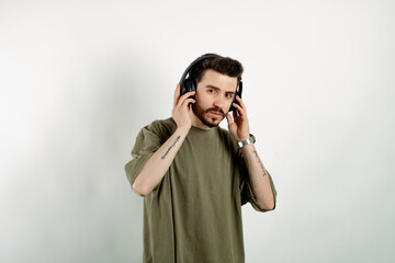 Wall Mural - Young man wearing khaki t-shirt posing isolated over white background wireless headphones meditating listening audio guide. Touching to headphone.