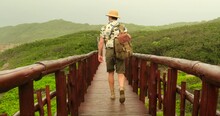 Male Traveler With A Backpack On His Back Walks Along A Wooden Bridge Among Tropical Grass. Guy In Safari Style, In A Hat And Shirt, Travel To Famous Places In The Amazon. Travel Blogger On Vacation 