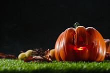 Against The Background Of A Black Gloomy Sky, A Pumpkin With A Carved Face And Illuminated From The Inside By A Candle, Autumn Fruits On Green Grass. Halloween Background.