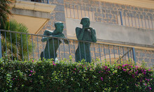 Two Statues Standing On Balcony  Leaning Against Handrails. 