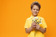 © Tatiana - a funny, curly-haired boy of school age stands in a yellow polo and examines a bouquet of daisies in his hands. Horizontal studio photography on orange background with empty space