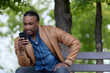 © Aleksandr - An African-American fashionable student enthusiastically reads an e-book on his smartphone while relaxing in the park. An interesting e-book is read by a black student sitting on a bench on his phone