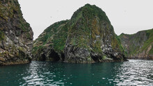 Rocky Islands In The Pacific Ocean. Seabirds Nest On Steep Slopes Covered With Sparse Vegetation. The Entrances To Dark Caves Are Visible Above The Water. Kamchatka. Starichkov Island