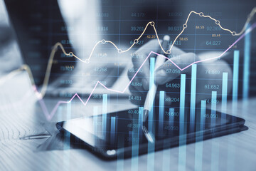 Poster - businessman hand using tablet and laptop at desktop with abstract glowing business chart with index and growth on blurry background. Finance, trade and market report concept. Double exposure.
