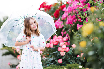 Wall Mural - Happy cute kid child girl with long blonde hair holding transparent umbrella posing over rose flowers in park outdoor. Cheerful little toddler at city street wear romantic style dress. Summer rain