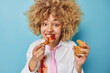 Beautiful cheerful woman feels very hungry eats fried nuggets with ketchup enjoys cheat meal wears formal clothing poses against blue background. Unhealthy nutrition and binge eating concept