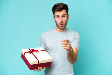 Wall Mural - Young caucasian man holding a gift isolated on blue background surprised and pointing front