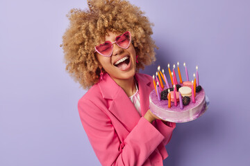 Wall Mural - Overjoyed woman with curly hair laughs out happily wears trendy heart shaped sunglasses and formal jacket holds big tasty cake celebrates anniversary enjoys party time isolated over purple background