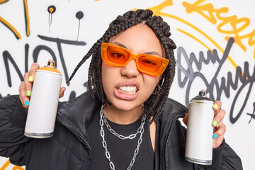 Poster - Female street artist with white teeth frowns face has braided hairstyle wears orange sunglasses black clothes and metal chains holds two aerosol sprays paints colorful graffiti on generic wall