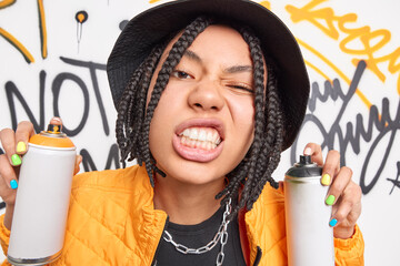 Sticker - Close up shot of female teenager holds two spray cans for drawing graffiti clenches teeth has cheeky expression braided hairstyle wears black hat and orange jacket poses against graffiti wall