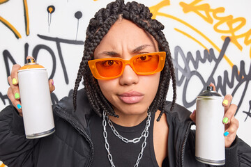 Poster - Displeased female grafitti artist holds two cans with paint creats street art wears trendy orange sunglasses and black jacket poses against colorful drawn wall. Street style and adolescence concept