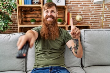 Sticker - Redhead man with long beard holding television remote control smiling with an idea or question pointing finger with happy face, number one