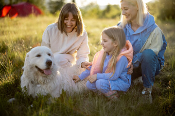 Portrait of two women with little girl and white dog sit together on nature during sunset. Homosexual relationship, parenting kid and travel in mountains concept