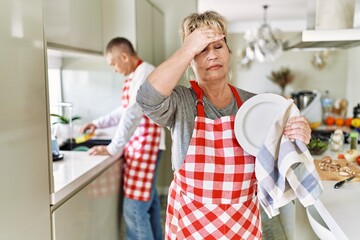 Poster - Middle age caucasian couple wearing apron washing dishes at home stressed and frustrated with hand on head, surprised and angry face