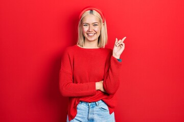 Canvas Print - Beautiful blonde woman wearing casual red sweater with a big smile on face, pointing with hand and finger to the side looking at the camera.