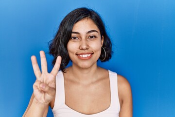 Sticker - Young hispanic woman standing over blue background showing and pointing up with fingers number three while smiling confident and happy.