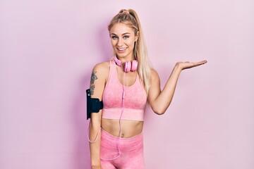 Wall Mural - Young beautiful caucasian woman wearing gym clothes and using headphones smiling cheerful presenting and pointing with palm of hand looking at the camera.