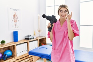 Poster - Young physiotherapist woman holding therapy massage gun at wellness center surprised with an idea or question pointing finger with happy face, number one