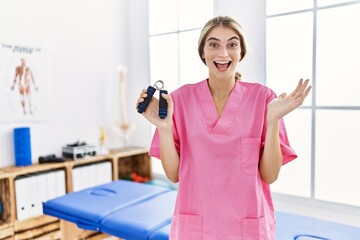 Poster - Young blonde woman working at pain recovery clinic holding hand strengthener celebrating achievement with happy smile and winner expression with raised hand