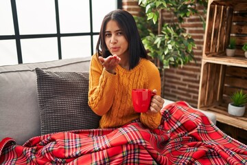 Poster - Young hispanic woman sitting on the sofa drinking a coffee at home looking at the camera blowing a kiss with hand on air being lovely and sexy. love expression.