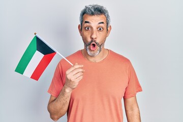 Sticker - Handsome middle age man with grey hair holding kuwait flag scared and amazed with open mouth for surprise, disbelief face