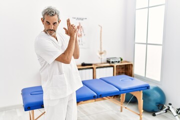 Canvas Print - Middle age hispanic therapist man working at pain recovery clinic holding symbolic gun with hand gesture, playing killing shooting weapons, angry face