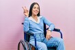 Beautiful woman with blue eyes sitting on wheelchair smiling looking to the camera showing fingers doing victory sign. number two.