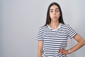 Wall Mural - Young brunette woman wearing striped t shirt puffing cheeks with funny face. mouth inflated with air, crazy expression.