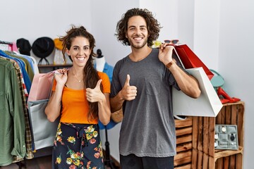 Wall Mural - Young hispanic couple holding shopping bags at retail shop doing happy thumbs up gesture with hand. approving expression looking at the camera showing success.