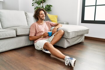 Sticker - Young hispanic man drinking coffee sitting on the floor at home.