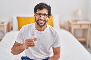 Sticker - Young hispanic man drinking glass of water sitting on bed at bedroom