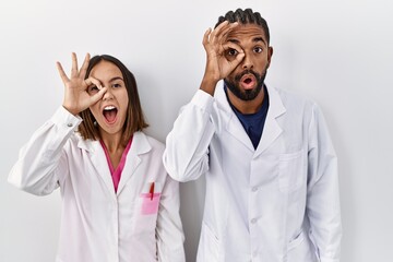 Canvas Print - Young hispanic doctors standing over white background doing ok gesture shocked with surprised face, eye looking through fingers. unbelieving expression.