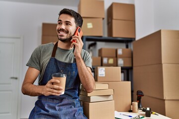 Wall Mural - Young hispanic man business worker talking on the smartphone drinking coffee at storehouse