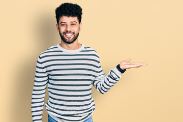 Sticker - Young arab man with beard wearing casual striped sweater smiling cheerful presenting and pointing with palm of hand looking at the camera.